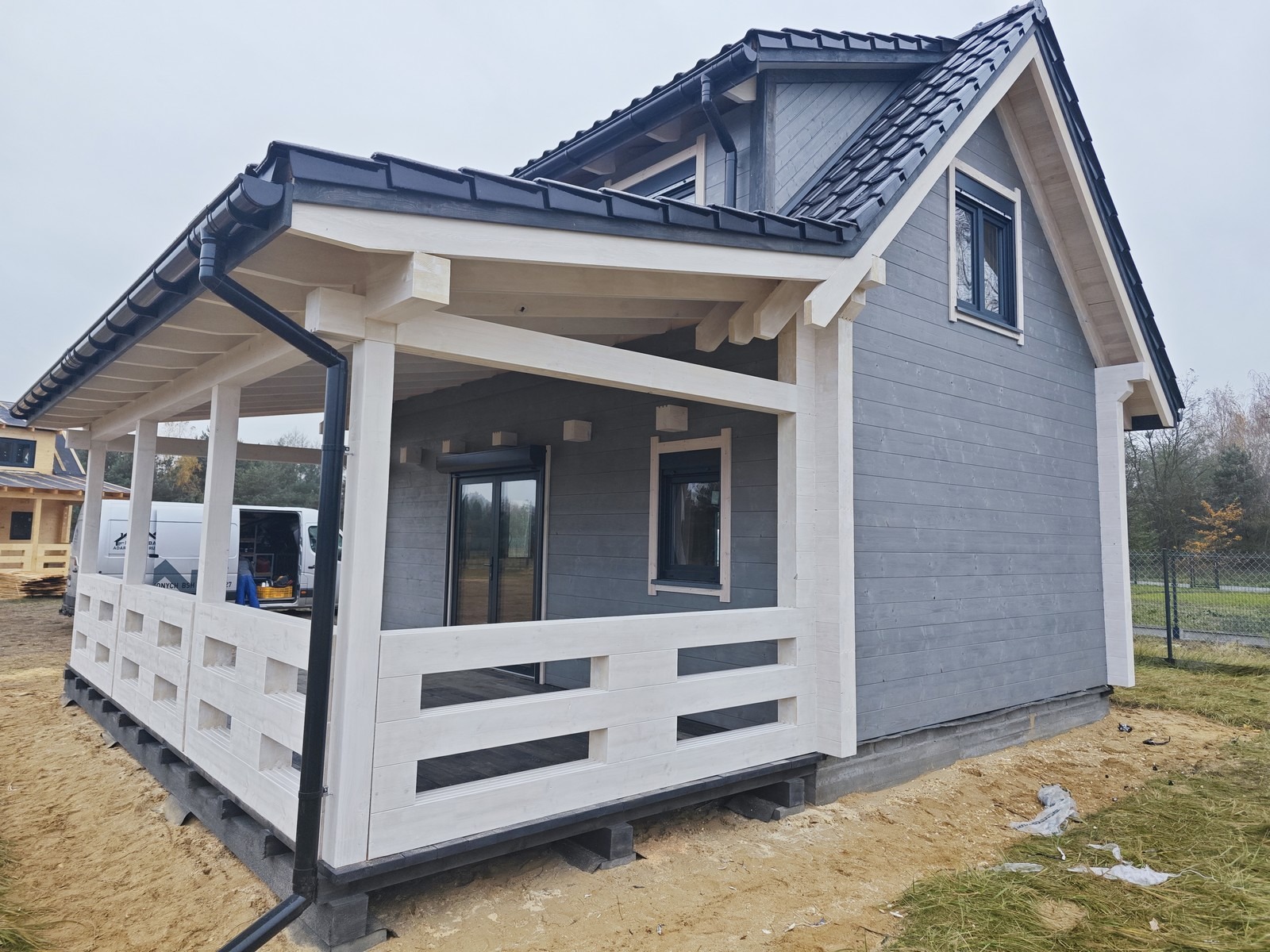 Szary dom z bali z białymi elementami wykończeniowymi, czarną dachówką i zadaszonym tarasem, widok z boku na etapie wykończenia terenu.