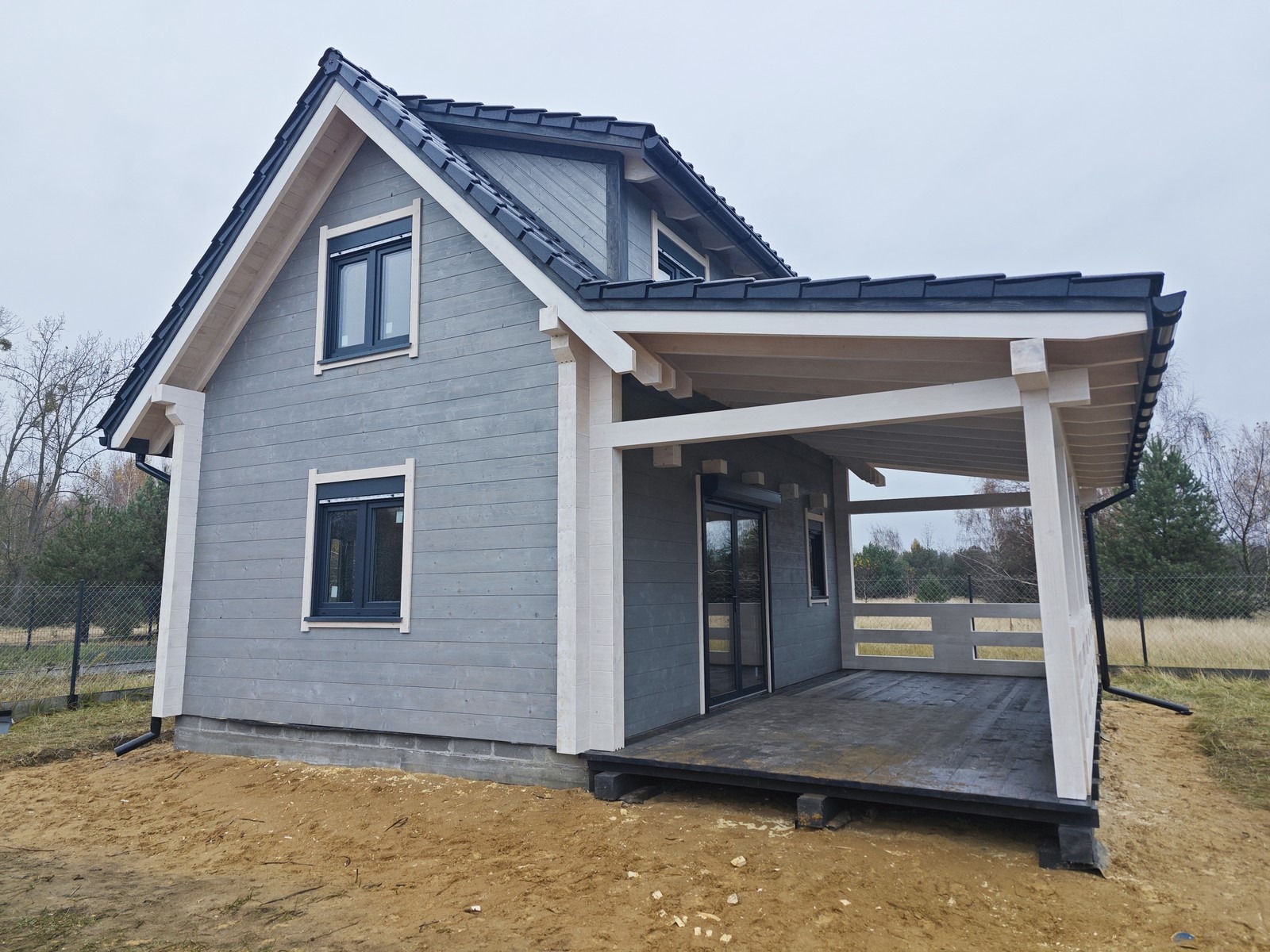 Szary dom z drewna z ciemną dachówką i zadaszonym tarasem, widok z zewnątrz na tle jesiennego krajobrazu.