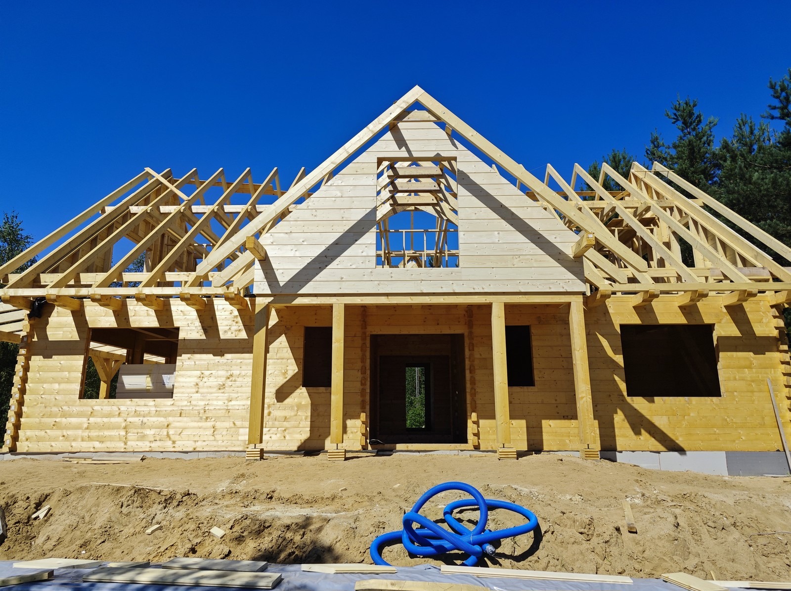 Surowa konstrukcja domu z bali w trakcie budowy, widoczne drewniane ściany, więźba dachowa i niebieski wąż do instalacji na ziemi.