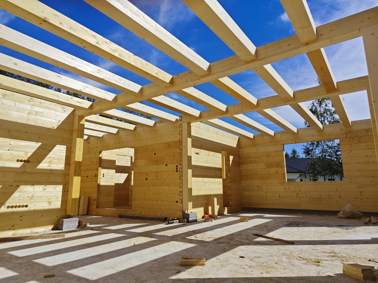 Surowy szkielet domu z bali w trakcie budowy, widoczne drewniane ściany, strop i niebieskie niebo przez otwory w dachu.