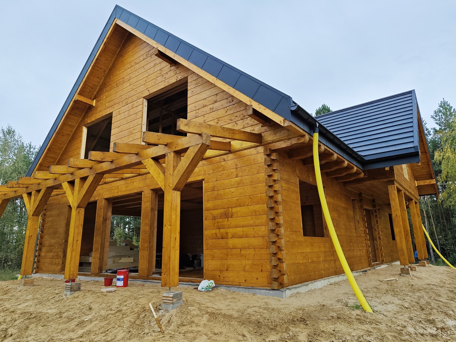 Surowy stan budowy domu z bali z ciemną blachodachówką, widoczne elementy konstrukcyjne i żółty wąż doprowadzający prawdopodobnie wodę, teren wokół domu piaszczysty.