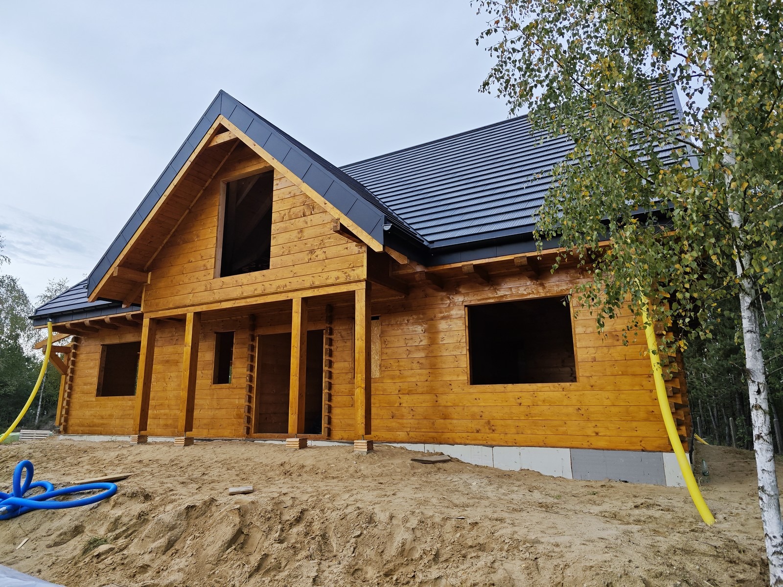Surowy drewniany dom z bali z czarnym dachem w trakcie budowy, widoczne instalacje i piasek.