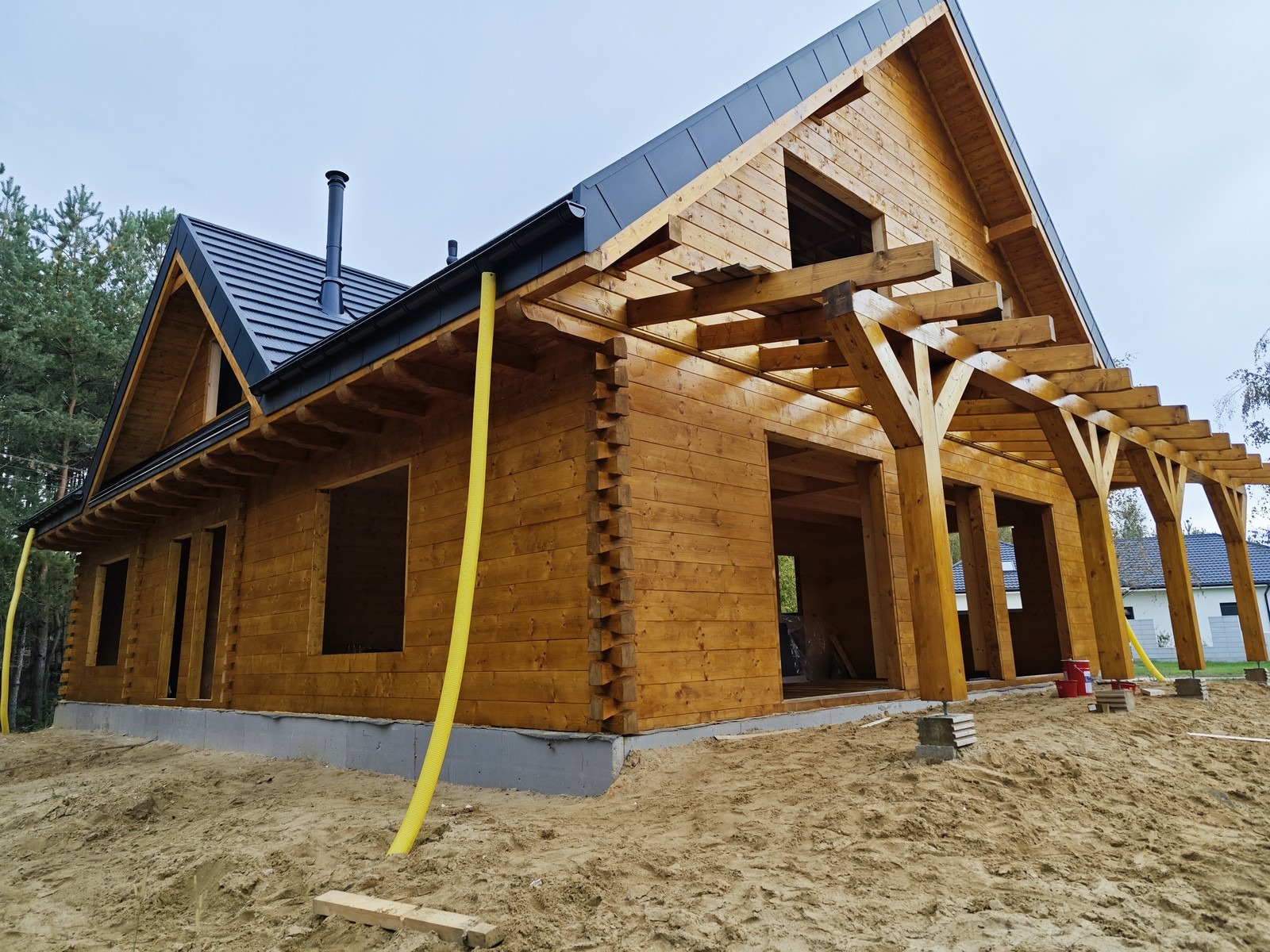 Surowy drewniany dom z bali w trakcie budowy, z widoczną konstrukcją dachu i zadaszeniem tarasu, na piaszczystym terenie, z żółtymi rurami wentylacyjnymi wystającymi z fundamentów.