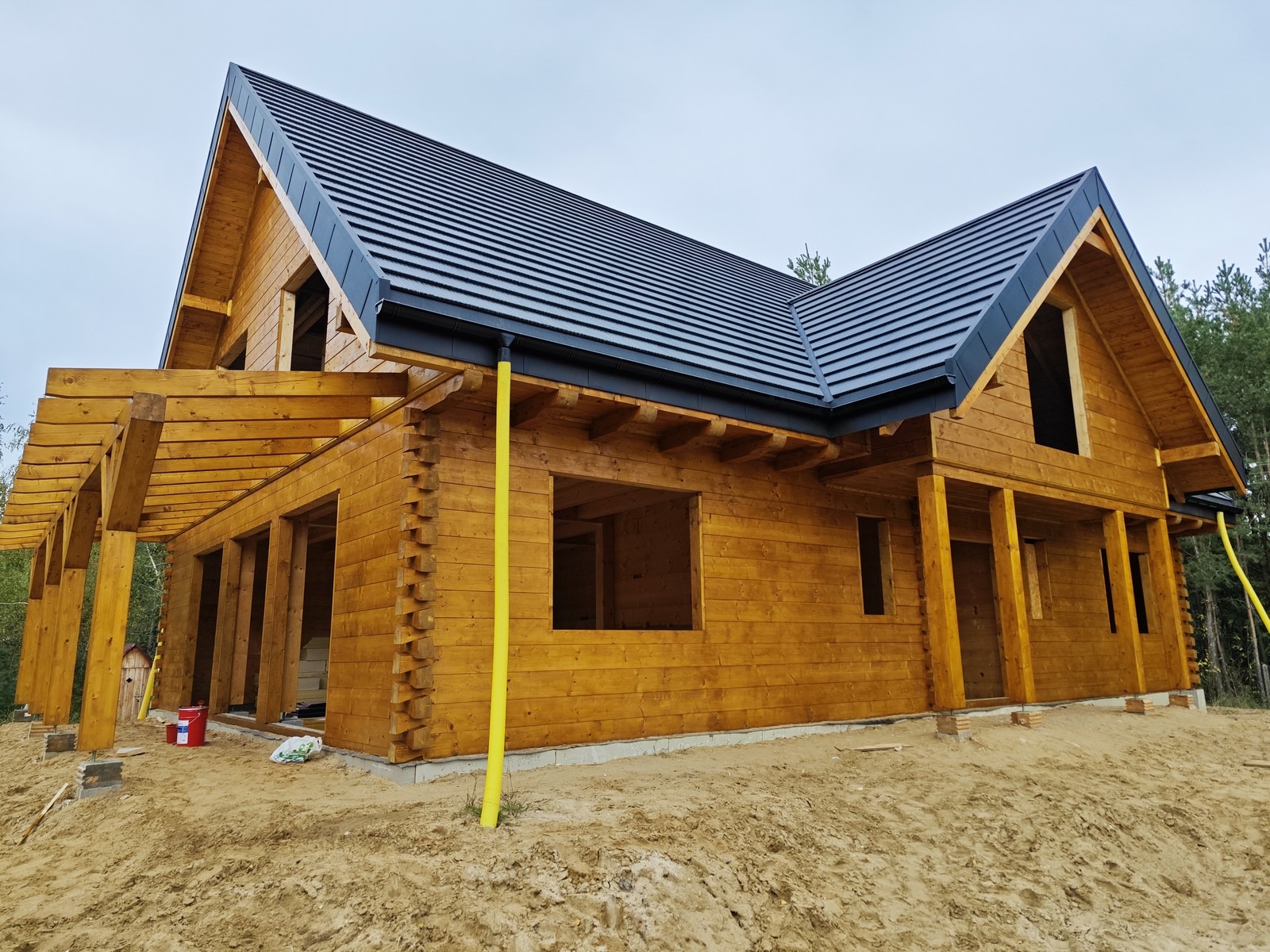 Surowy, dwukondygnacyjny dom z bali z czarnym dachem i żółtą rurą spustową, w trakcie budowy, widoczne niezabudowane otwory okienne i drzwiowe, teren wokół domu piaszczysty, w tle las.