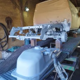 Podwozie samochodu po piaskowaniu i zabezpieczeniu antykorozyjnym, widoczny zbiornik paliwa, całość umieszczona na metalowej konstrukcji w warsztacie.