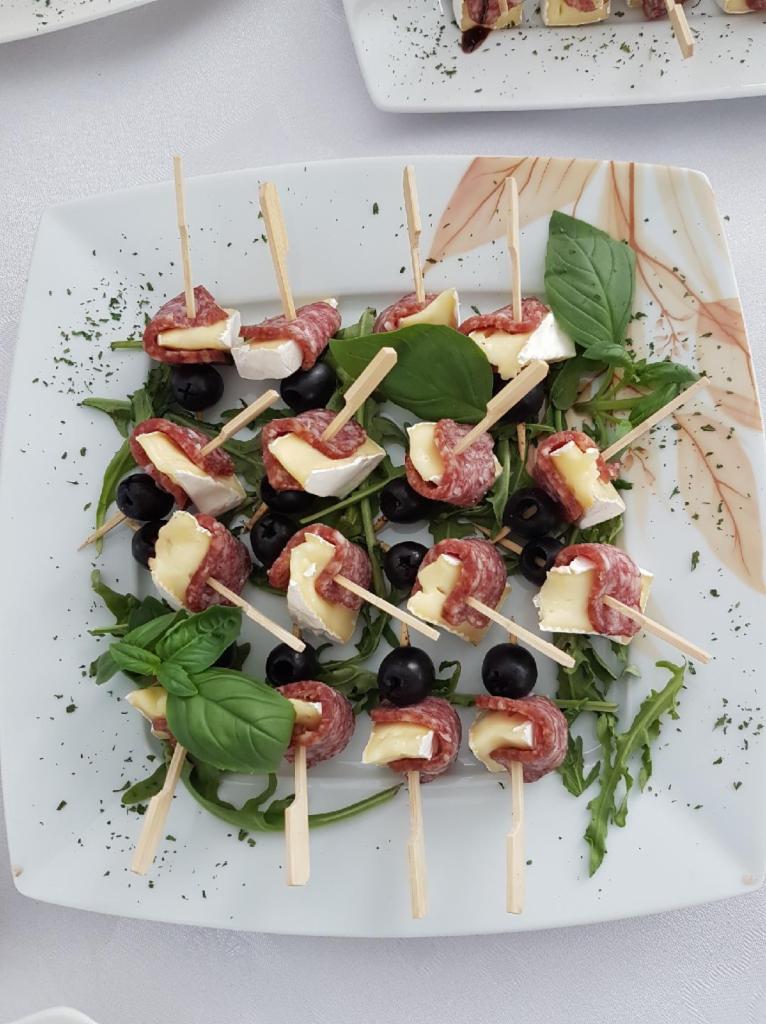 Elegancko podane koreczki: salami, ser brie i oliwki na wykałaczkach, ułożone na rukoli i udekorowane listkami bazylii, na kwadratowym talerzu z delikatnym wzorem.