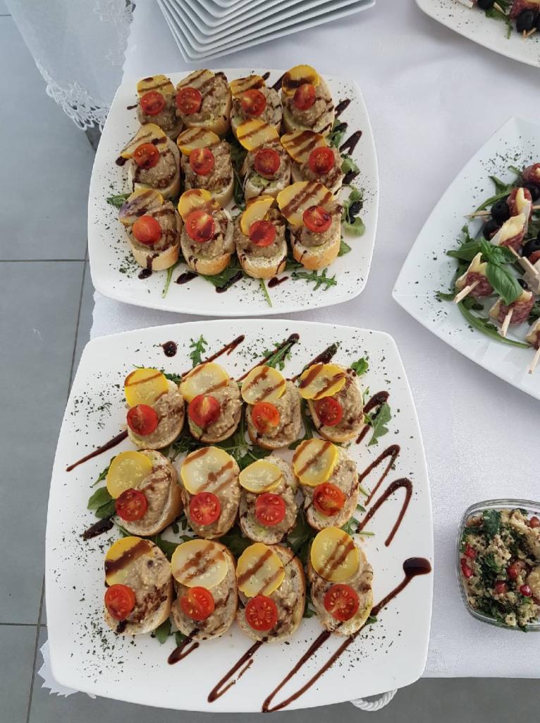 Dwie kwadratowe białe tace z kanapkami na bagietkach z pastą, pomidorkiem cherry i plasterkiem marynowanej cukinii, polane sosem balsamicznym, na stole z białym obrusem.