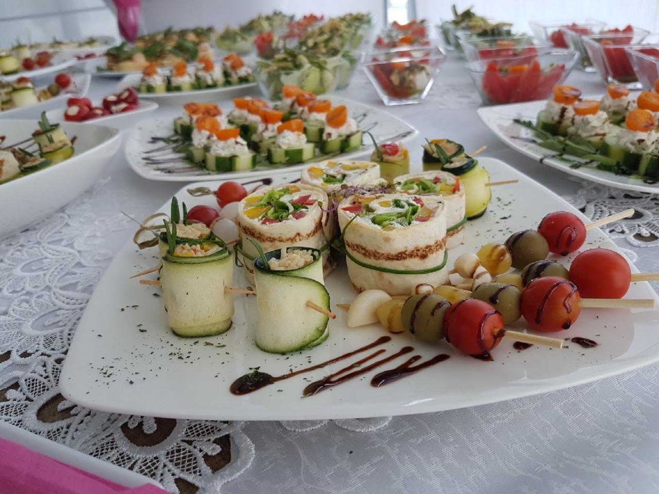 Dekoracyjnie podane przekąski: roladki z tortilli i warzyw, koreczki z pomidorków koktajlowych i oliwek na białym talerzu z sosem balsamicznym, w tle inne talerze z cateringiem.