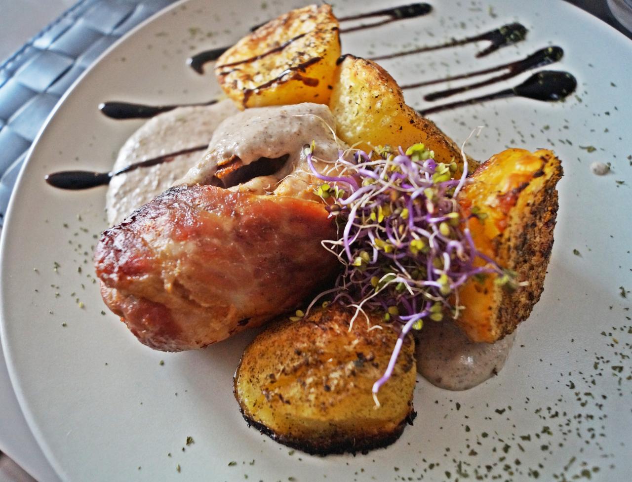 Elegancko podane danie: pieczone ziemniaki, mięso w szynce, kremowy sos i świeże kiełki na białym talerzu, ozdobione ciemnym sosem i ziołami.