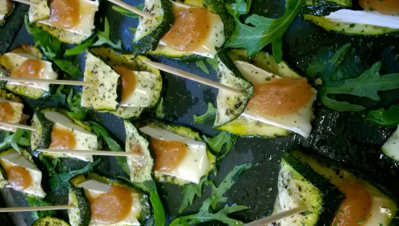 Koreczki z grillowanej cukinii, sera brie i pomarańczowego sosu na ciemnym, kamiennym talerzu, udekorowane świeżą rukolą i spięte wykałaczkami.