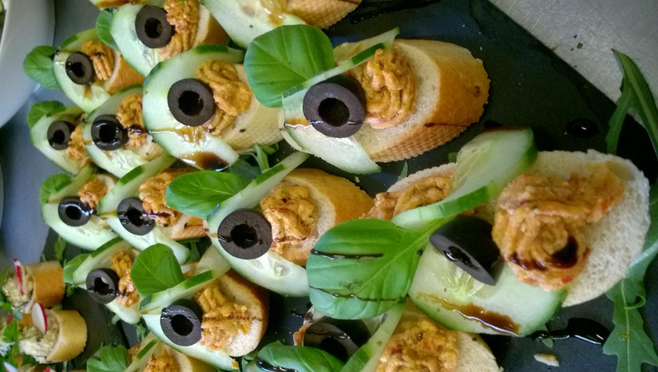 Dekoracyjne kanapeczki z pastą, oliwką i ogórkiem, polane sosem balsamicznym, ułożone rzędami na ciemnym talerzu.