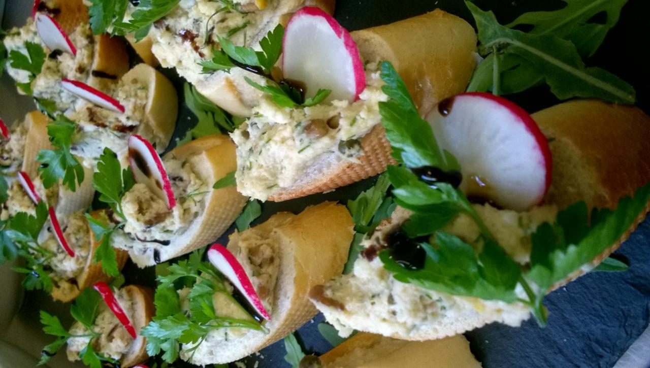 Apetyczne kanapeczki na bagietce z pastą, rzodkiewką, natką pietruszki i sosem balsamicznym, ułożone na ciemnym talerzu z rukolą.