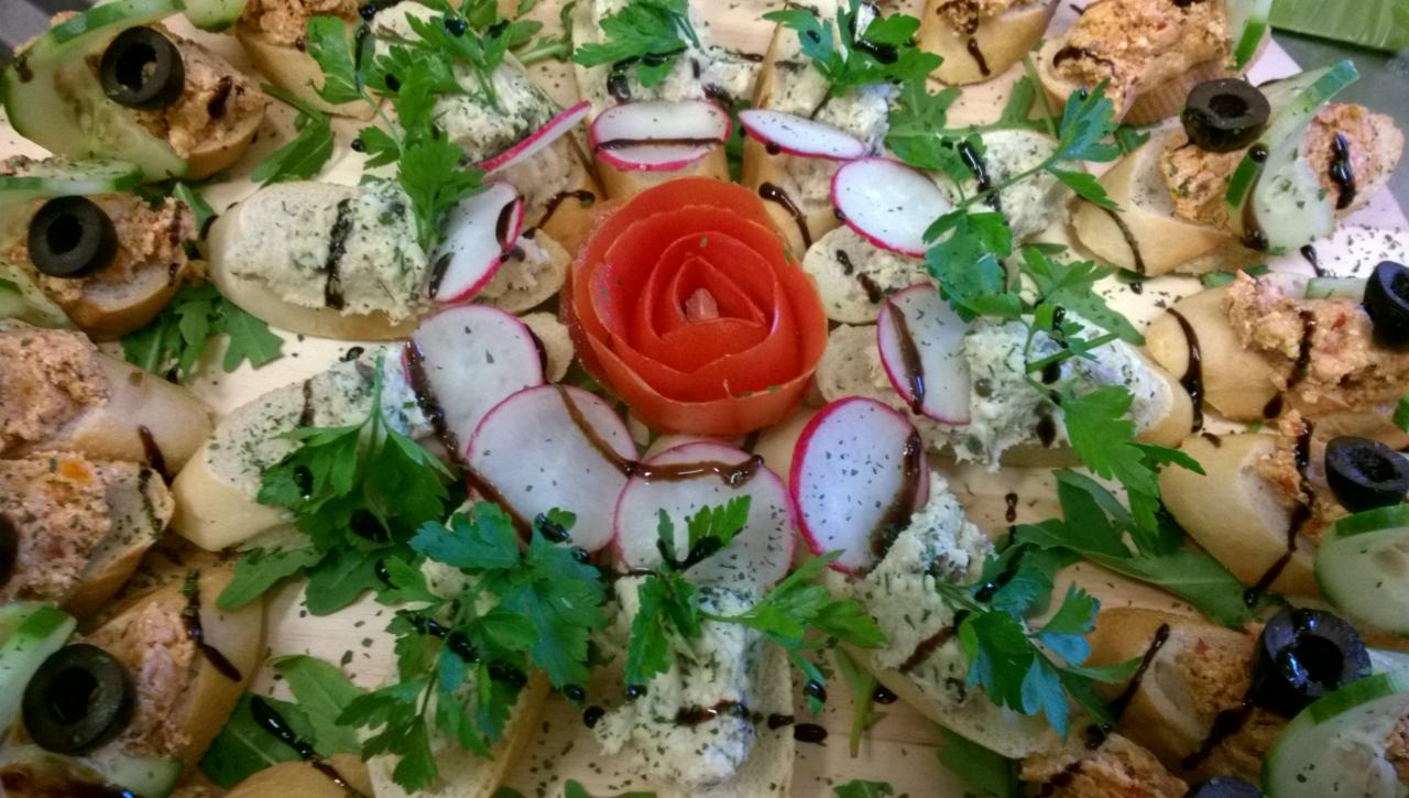 Dekoracyjna kompozycja kanapek z różnymi pastami, rzodkiewką, ogórkiem, oliwkami i sosem balsamicznym, ułożonych na drewnianej desce, z centralnie umieszczoną różą z pomidora.