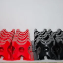 Rząd czerwonych i czarnych, wydrukowanych w 3D, plastikowych uchwyt&oacute;w na kable z otworem, ułożonych na białym tle.