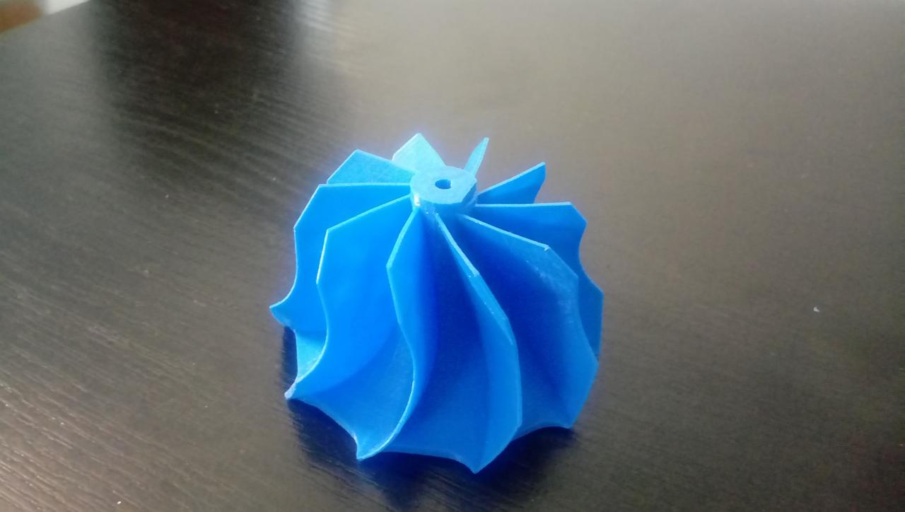 Niebieski model turbiny wydrukowany w 3D, widoczny na ciemnym, drewnianym blacie. Model ma spiralne łopatki i otwór w górnej części.
