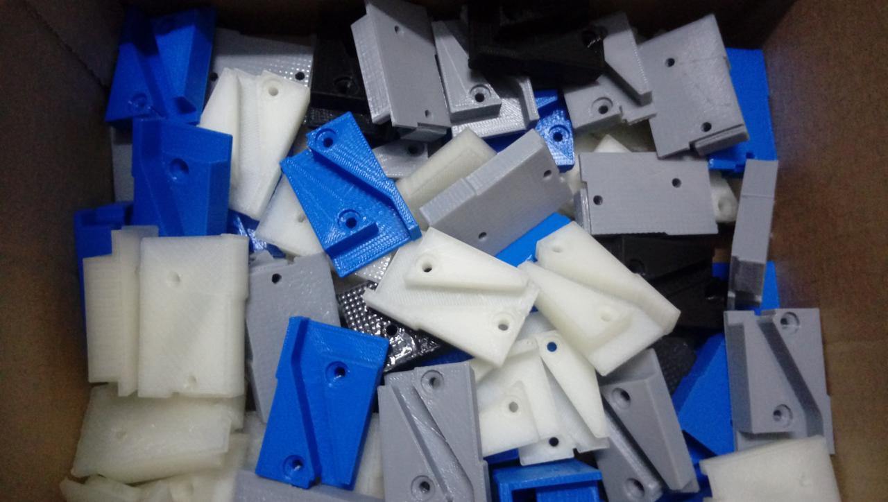 Karton wypełniony różnokolorowymi (białe, szare, niebieskie, czarne) elementami z tworzywa sztucznego z widocznymi otworami, prawdopodobnie wyprodukowanymi za pomocą drukarki 3D.