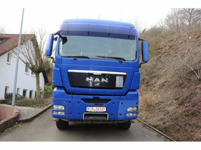 Niebieski ciągnik siodłowy MAN sfotografowany z przodu na tle budynku i wzgórza z drzewami.