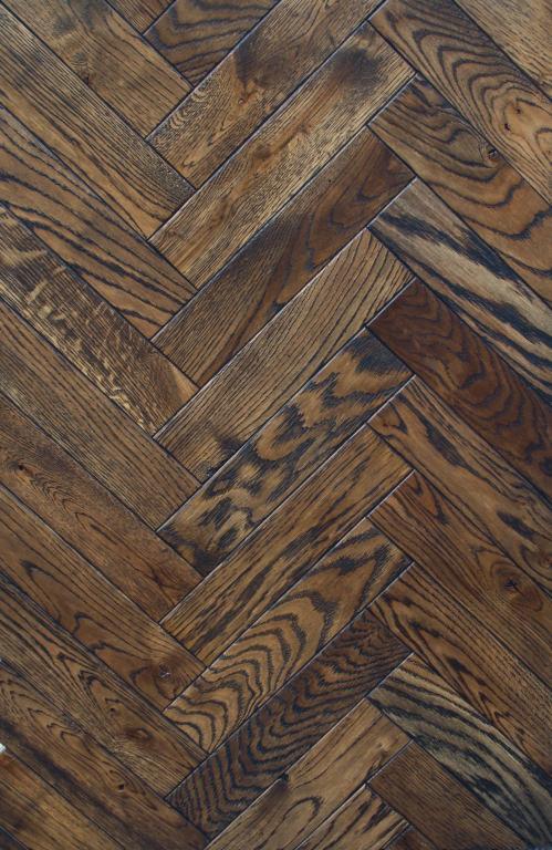 Ciemny, lakierowany parkiet ułożony we wzór jodełki francuskiej, z wyraźnym usłojeniem i zróżnicowaniem kolorystycznym poszczególnych klepek.