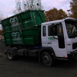 Biała ciężarówka z dwoma zielonymi kontenerami na gruz, z logo firmy 'Green' i numerem telefonu, stojąca na placu budowy z widocznymi resztkami gruzu w tle, pochmurne niebo.