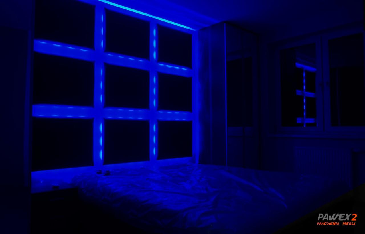Nastrojowe oświetlenie LED w pokoju, niebieskie światło podkreśla geometryczny wzór na ścianie nad łóżkiem, widoczny fragment szafy i okna w tle.