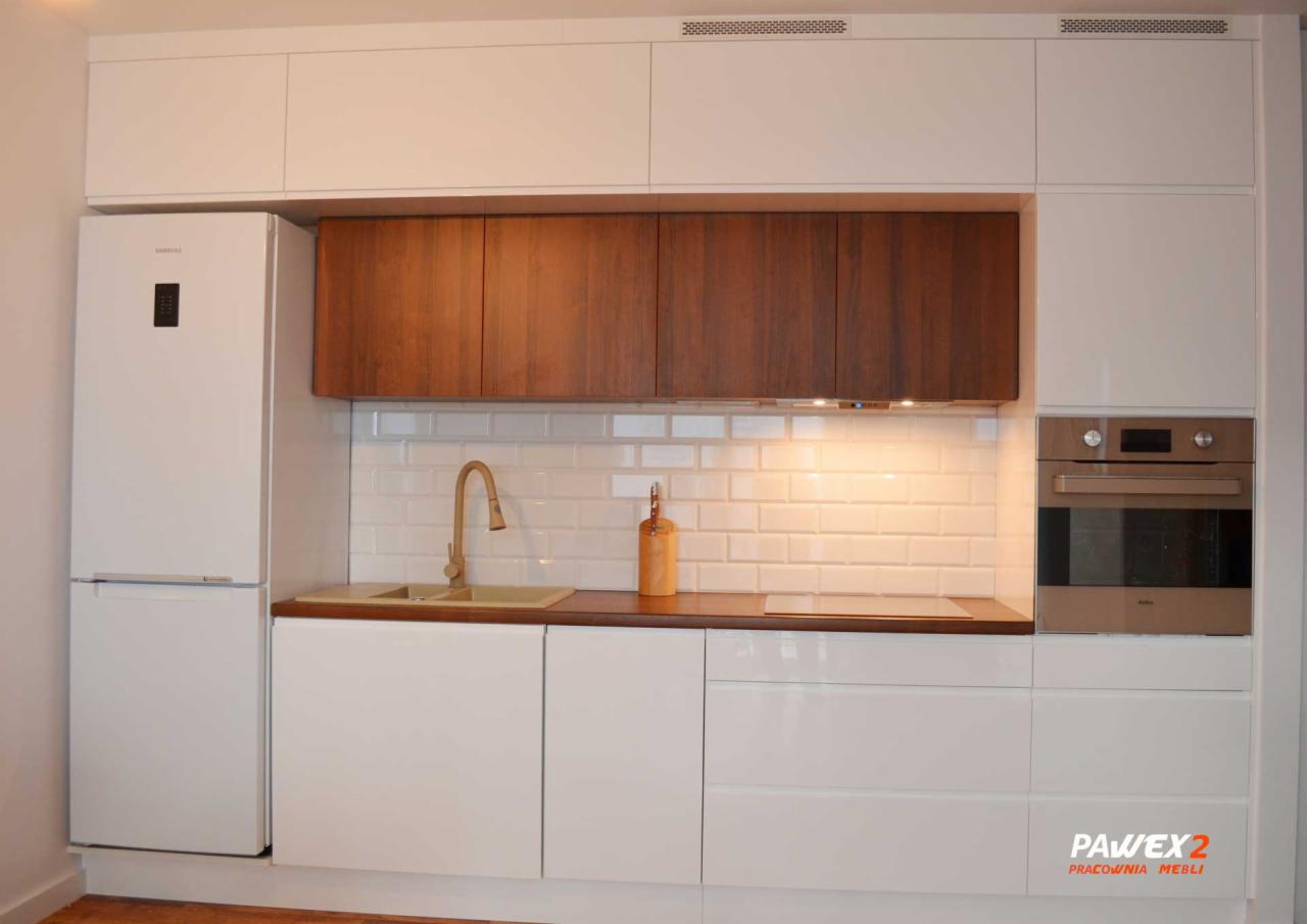 Nowoczesna kuchnia z białymi szafkami, drewnianymi blatami i złotą armaturą. Widoczny piekarnik w kolorze miedzianym i biała lodówka Samsung. Na blacie dekoracyjna butelka.