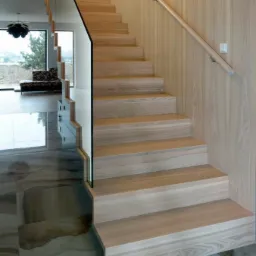 Drewniane schody z balustradą ze szkła i drewnianą poręczą, dopasowane kolorystycznie do okładziny ściennej, na marmurowej podłodze w nowoczesnym wnętrzu.