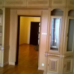 Klasyczna zabudowa drewniana w salonie z parkietem, obejmująca szafki, p&oacute;łki i obudowę drzwi, w odcieniach jasnego drewna z ciemniejszymi akcentami, widok na wnętrze pokoju z ciemnymi drzwiami.