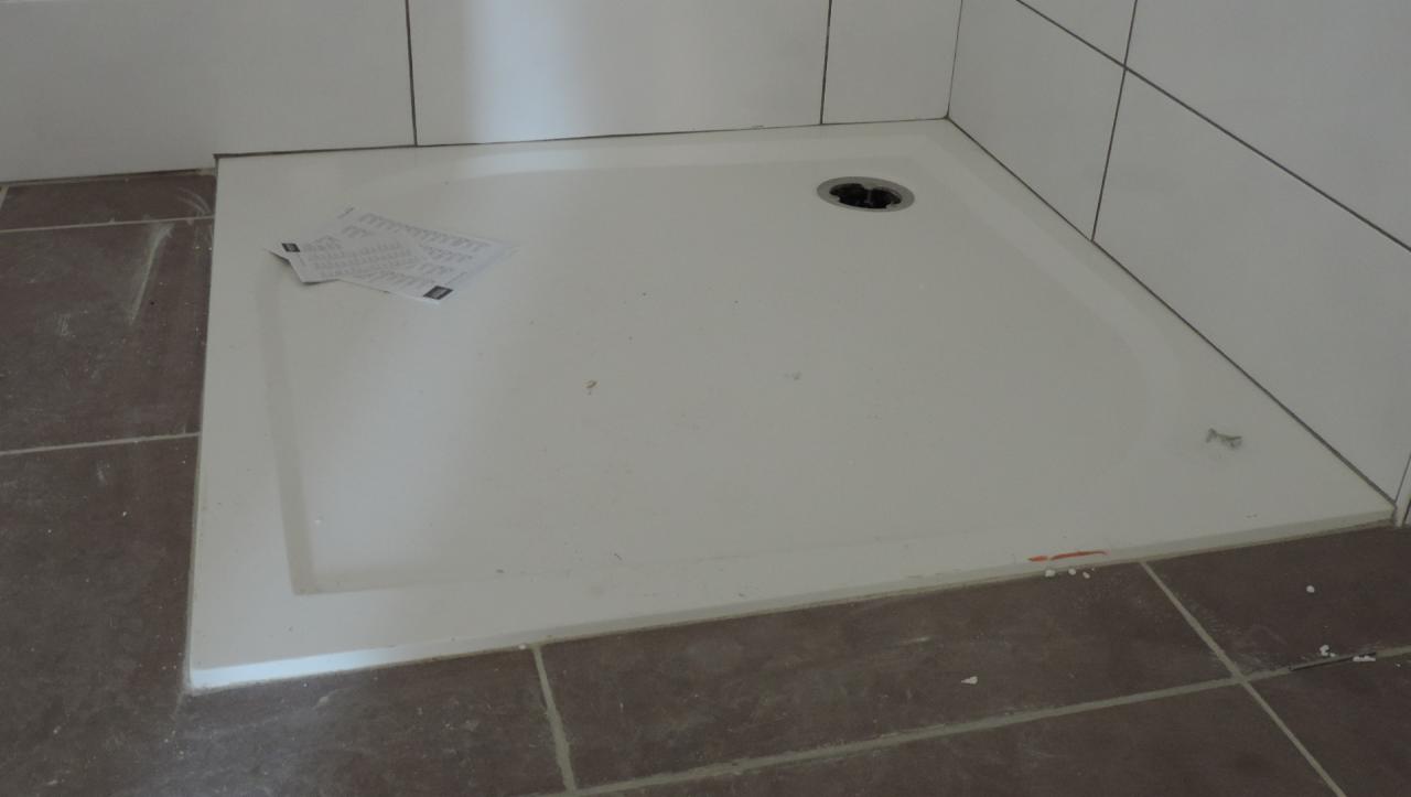 Biały brodzik w rogu łazienki, z widocznym odpływem i dokumentacją techniczną na powierzchni, otoczony brązowymi płytkami podłogowymi i białymi płytkami ściennymi.