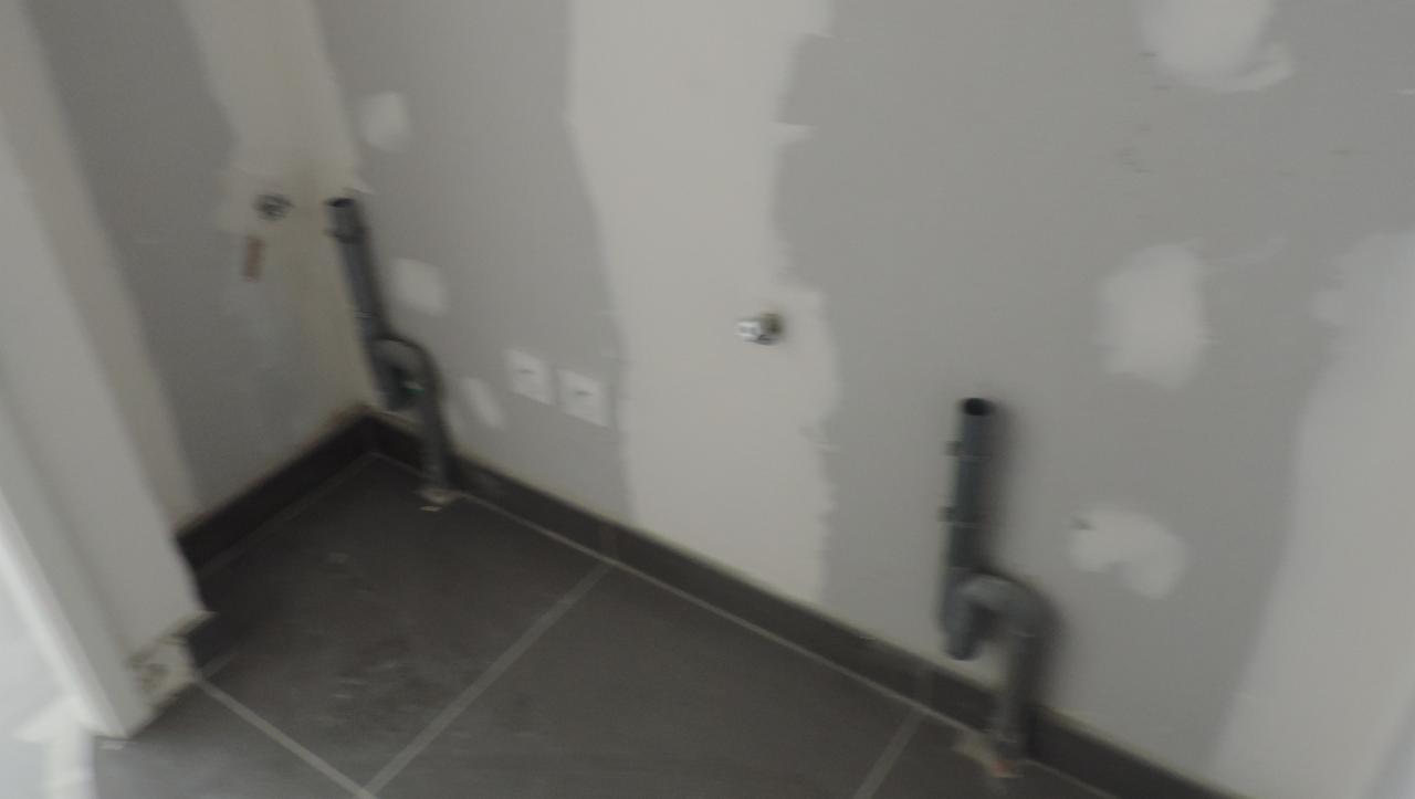 Surowe wykończenie łazienki w trakcie remontu: widoczne rury kanalizacyjne wystające ze ściany, przygotowane podłączenia wodne i szara, kwadratowa płytka podłogowa.