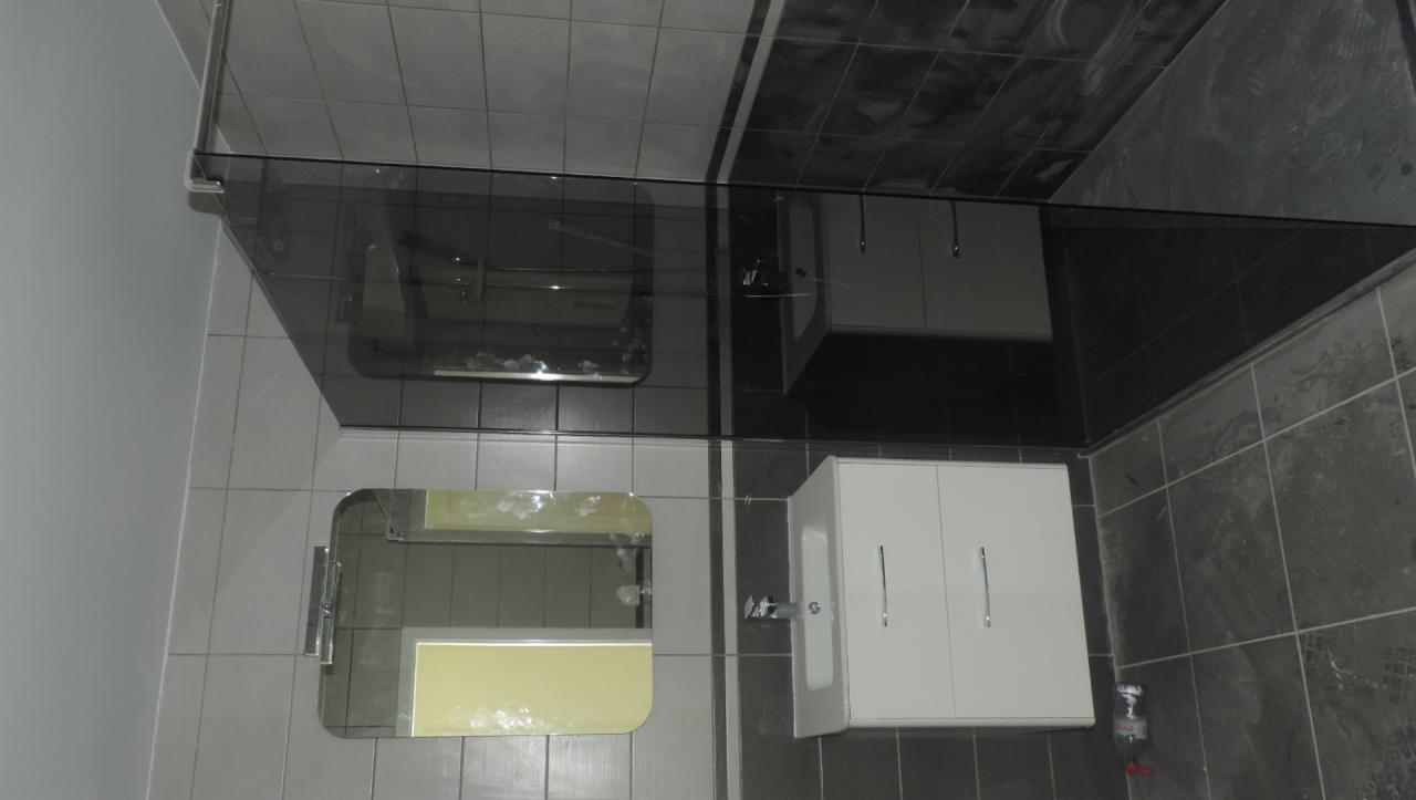 Wykończona łazienka z szarymi i czarnymi płytkami, białą szafką pod umywalką z chromowanymi uchwytami, oraz lustrem z żółtym akcentem. Widoczna szklana kabina prysznicowa.