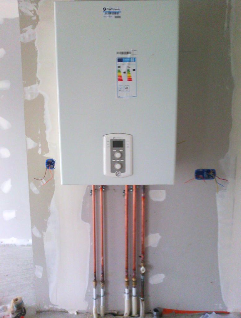 Biały kocioł gazowy z panelem sterowania, zamontowany na ścianie w trakcie prac instalacyjnych, widoczne miedziane rury i puszki elektryczne.