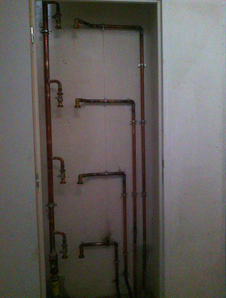 Instalacja rur miedzianych na ścianie wnęki, widoczne liczne złączki i zawory, częściowo zamontowane, w tle surowa ściana.