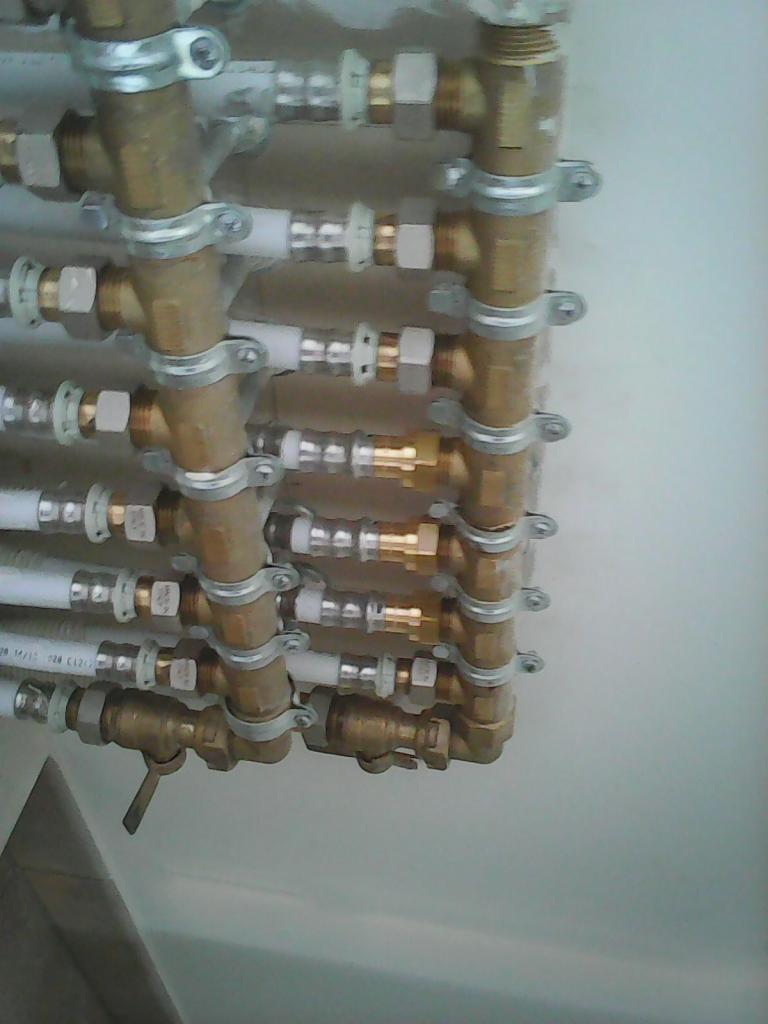 Rzędy mosiężnych i białych rur instalacyjnych, przymocowanych do ściany za pomocą metalowych obejm, tworzące rozdzielacz.