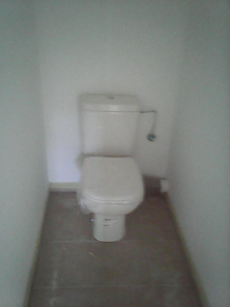 Nowa biała toaleta kompaktowa zamontowana w małej wnęce łazienki, widoczne szare płytki podłogowe i białe ściany.