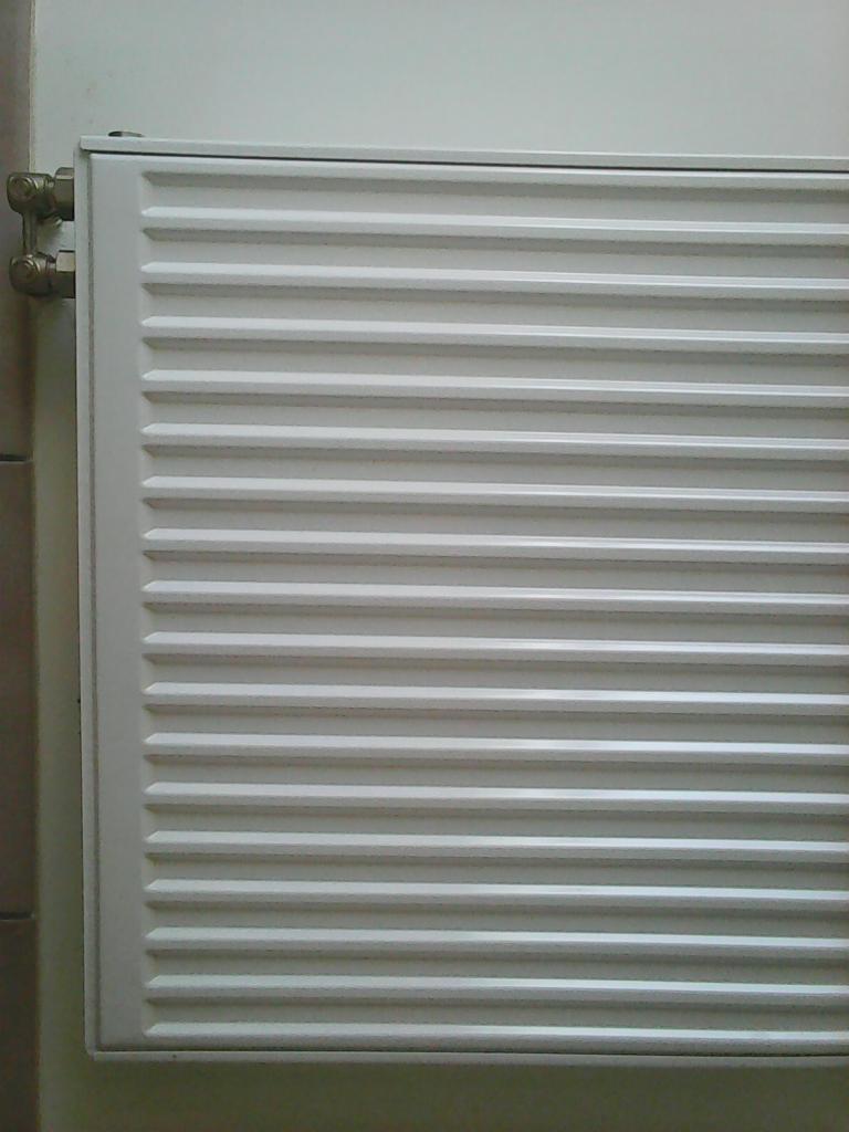 Biały, żebrowany grzejnik centralnego ogrzewania zamontowany na ścianie, widoczne mosiężne zawory przyłączeniowe.