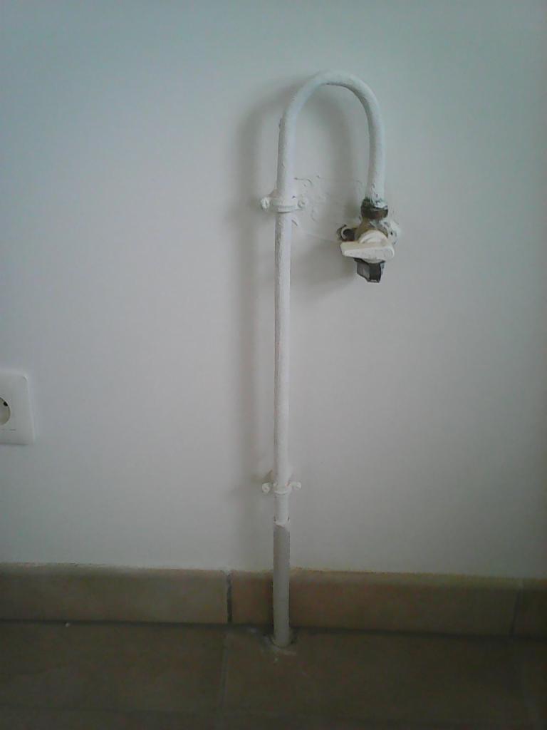 Biała rura gazowa z zaworem, pomalowana farbą, biegnąca wzdłuż ściany z gniazdkiem elektrycznym, na tle beżowej podłogi i listwy przypodłogowej.