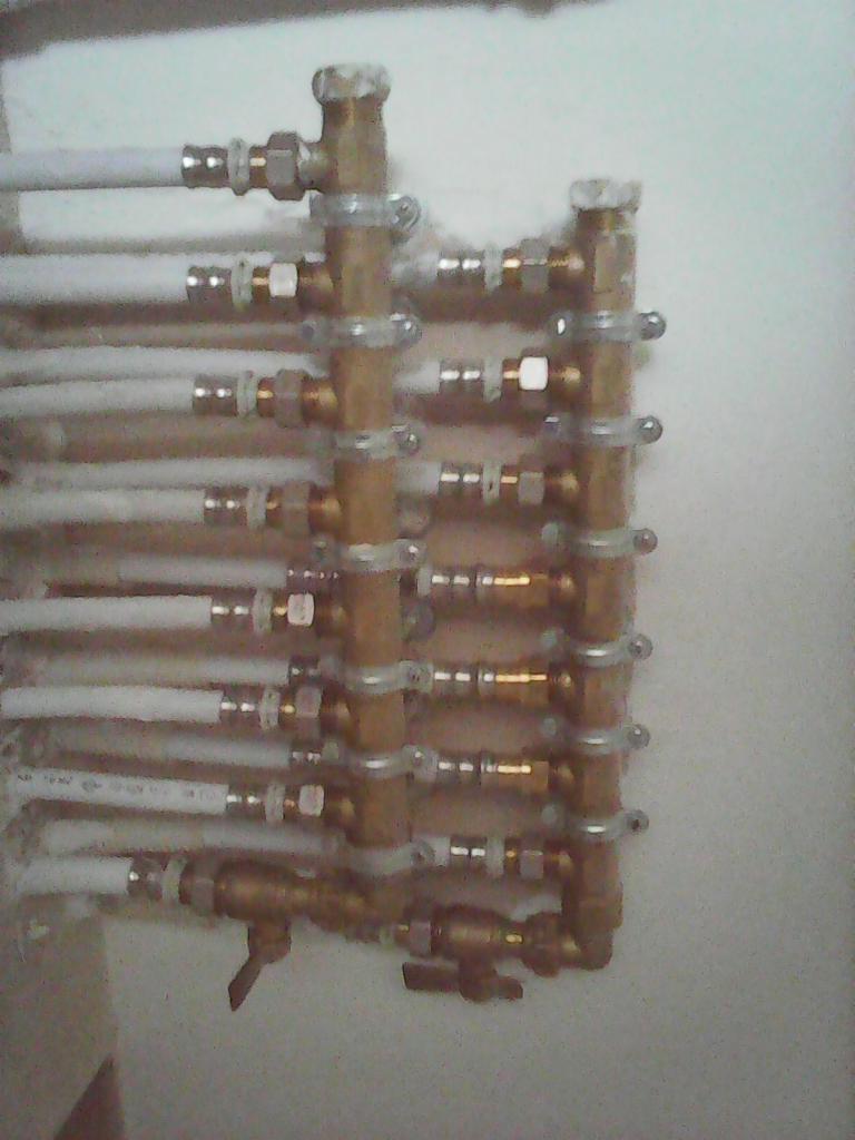 Szereg białych rur przymocowanych do ściany za pomocą metalowych uchwytów i połączonych mosiężnymi złączkami, tworzących pionowy kolektor instalacji grzewczej.