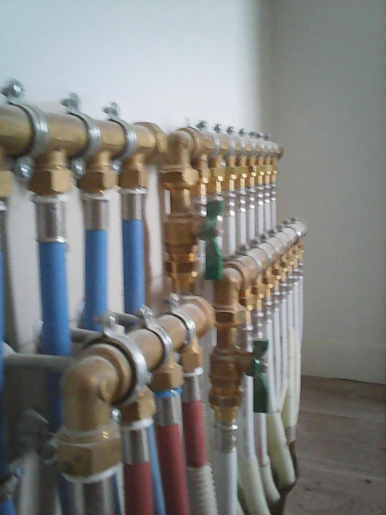 Rozdzielacz instalacji centralnego ogrzewania z rurami w kolorach niebieskim, czerwonym i białym, z zaworami kulowymi, zamocowany na ścianie.