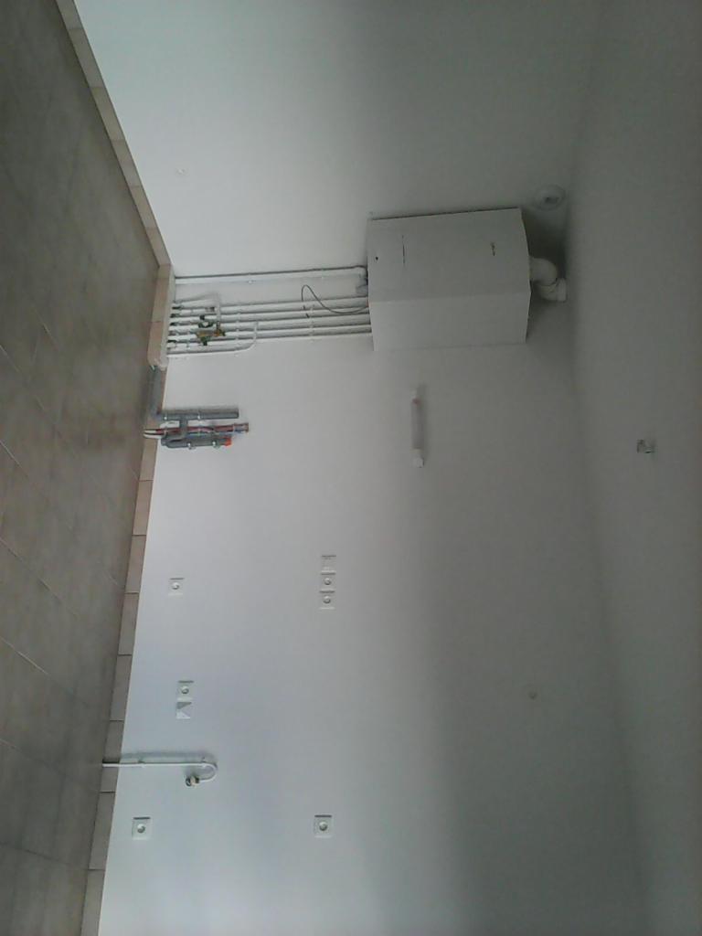 Instalacja grzewcza z rurami i białym kotłem zamontowana na ścianie w pomieszczeniu z szarą glazurą, widok z dołu.