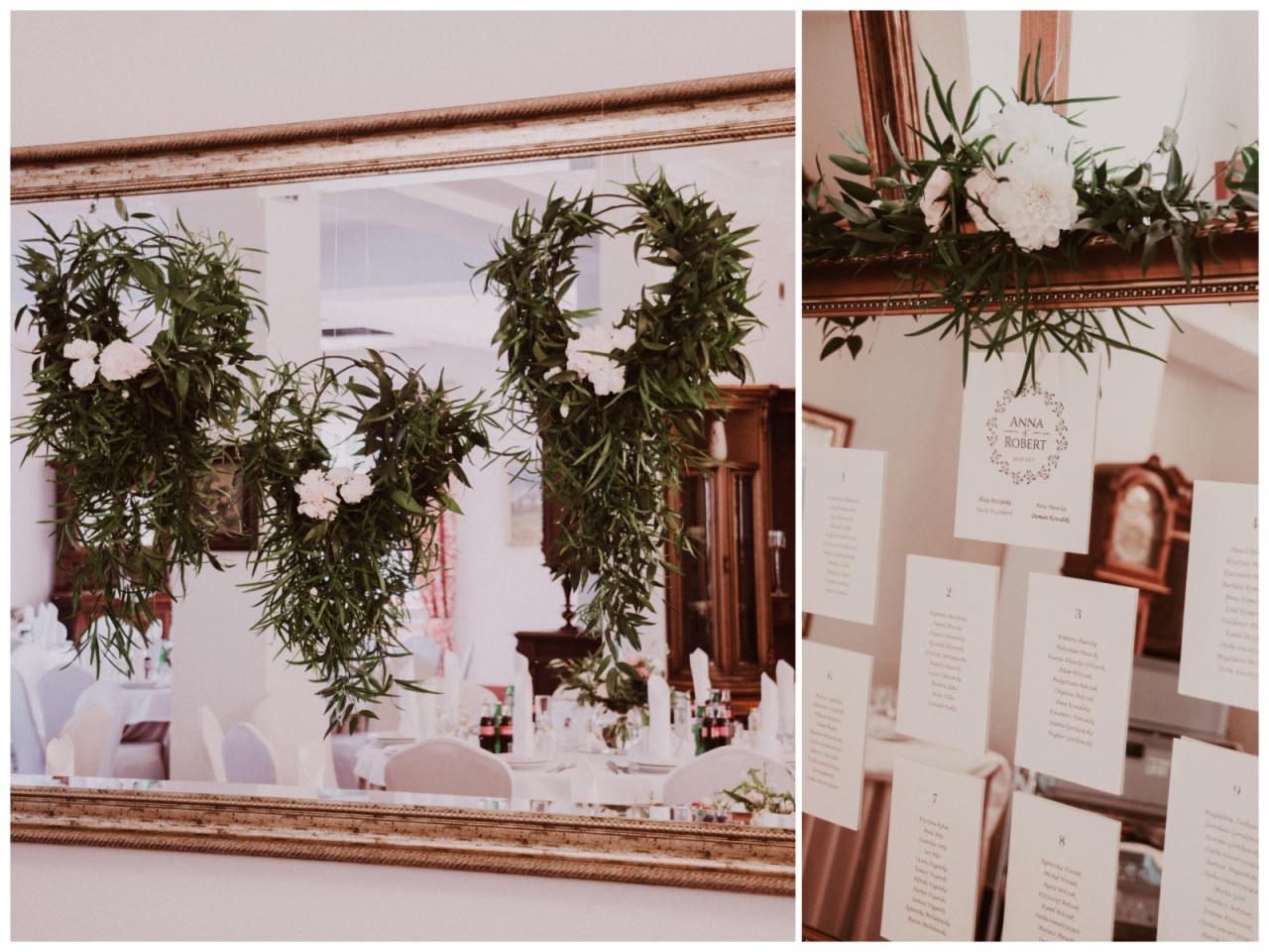 Dekoracja sali weselnej z lustrem w złotej ramie, na której zawieszone są kompozycje z zielonych liści i białych kwiatów, oraz kartki z numerami stołów i imionami gości.