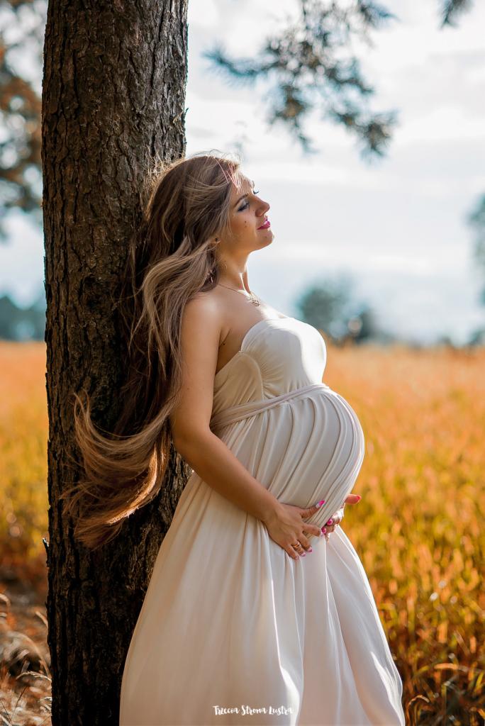 Kobieta w zaawansowanej ciąży, w zwiewnej sukni, opiera się o drzewo na tle pola w jesiennych barwach, z zamkniętymi oczami i uśmiechem na twarzy.