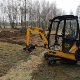 Żółta mini koparka JCB 8015 na błotnistym terenie, świeżo wykopany rów z ziemią w tle, drzewa bez liści i wyznaczone sznurkiem granice wykopu.