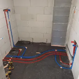 Wnętrze łazienki w trakcie remontu z widoczną instalacją ogrzewania podłogowego z rur w kolorze czerwonym i niebieskim, zamocowanych do ściany z betonu komórkowego, z dwoma wkrętarkami...