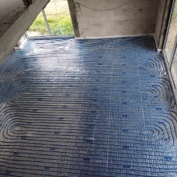 Montaż niebieskich rur ogrzewania podłogowego na folii w domu w stanie surowym w Mońkach, widok z góry, przy oknach.