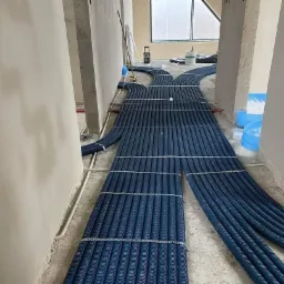 Wiązki niebieskich rur karbowanych ułożone równolegle na podłodze w surowym wnętrzu w trakcie instalacji systemu wentylacji lub rekuperacji, widoczne nieotynkowane ściany i okno w tle.