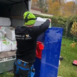 Pracownik w czarnej bluzie z logo firmy 'awterm.pl MANUFAKTURA CIEPŁA' w trakcie pracy z niebieską maszyną do aplikacji materiału izolacyjnego, widoczne worki z materiałem w przyczepie i żółty wąż.