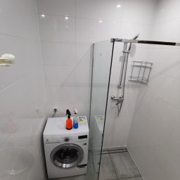 GKWORK JAKUB KLEBAŃSKI - Jasna łazienka po remoncie z kabiną prysznicową typu walk-in, płytkami w odcieniach bieli i szarości oraz pralką Electrolux z butelkami środków czyszczących na górze.