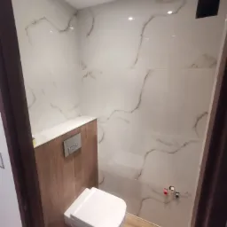 Nowoczesna, mała łazienka po remoncie: podwieszana toaleta z chromowanym przyciskiem spłukiwania, ściany wyłożone białymi płytkami imitującymi marmur z brązowymi żyłkami, drewnopodobne panele...