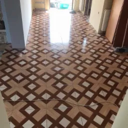 Nowo ułożona podłoga z płytek ceramicznych w geometryczny wz&oacute;r, brązowo-biała kolorystyka, widoczna drabina i otwarte drzwi na zewnątrz, przew&oacute;d elektryczny na podłodze.