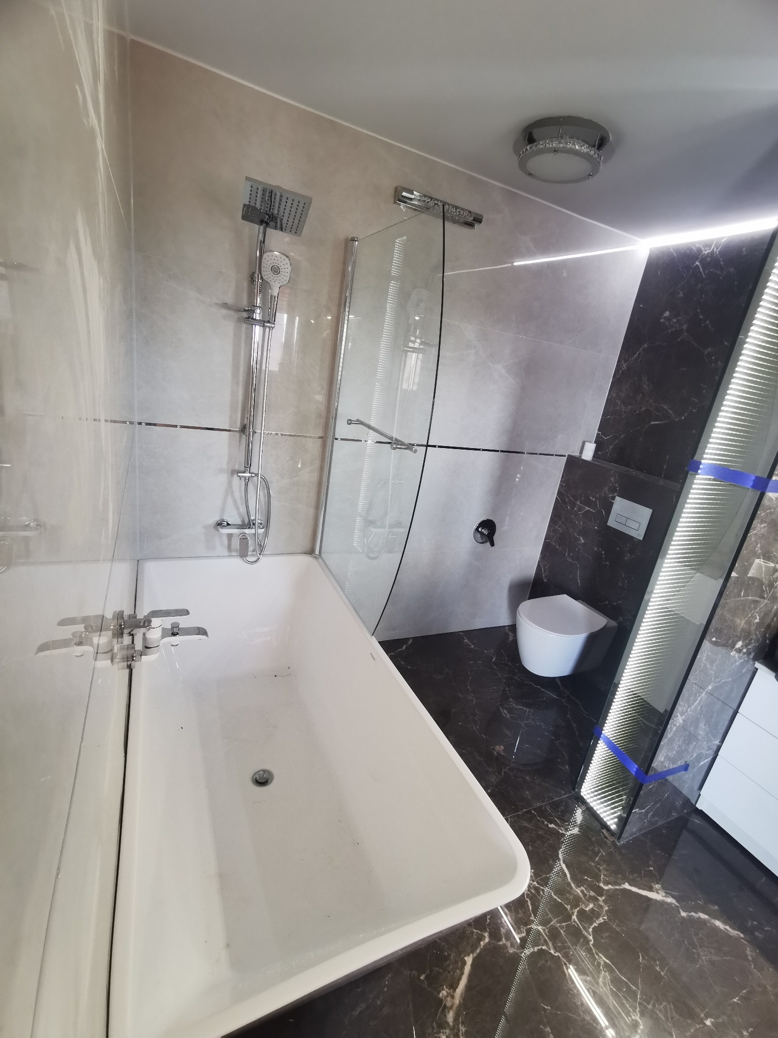 Nowoczesna łazienka z marmurową podłogą i ścianami, białą wanną z hydromasażem, szklanym panelem prysznicowym i podświetlaną wnęką, widok z góry na armaturę.